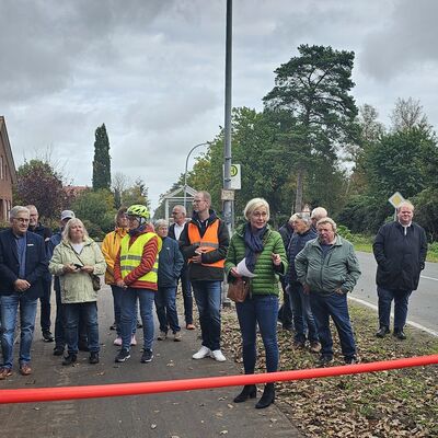 Das Bild zeigt eine Gruppe von Menschen auf dem neuen Radweg. Die Landr&auml;tin Karin Harms h&auml;lt eine Rede. In der Mitte des Bildes ist ein rotes Band zusehen.